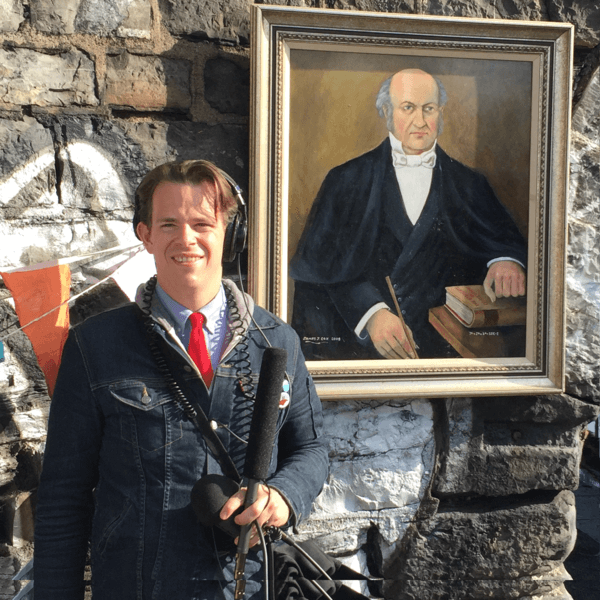 Picture of Sam holding a shotgun mic in front of a painting of William Rowan Hamilton on the side of Broom Bridge